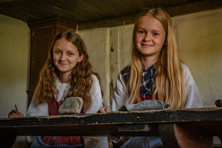 To smilende piger i tidstypiske dragter sidder på skolebænken i den gamle landsbyskole fra Hinge på Hjerl Hede.